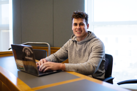 Burke Griffin '23 A male student sits in a classroom with his laptop in front of him, smiling at the camera