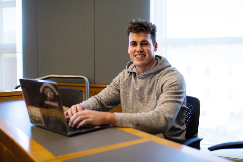 A male student sits in a classroom with his laptop in front of him, smiling at the camera