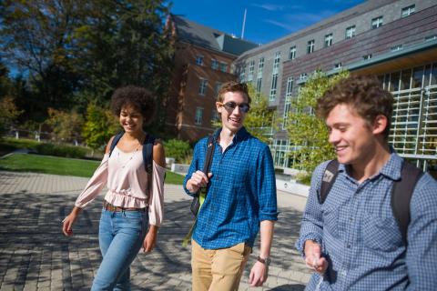 Three students stroll in the stone-paved courtyard, smiling with backpacks on. In the background, is grass and the exterior of the Paul College building.