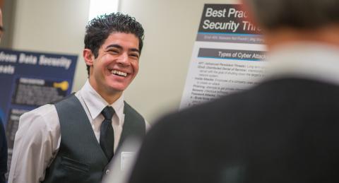 A professionaly dressed student smiles largely as he looks towards a professor in the foreground. He stands next to a large-scale poster showcasing research.