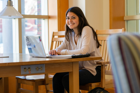 A female student sits at a table in the UNH library with her laptop and backpack, smiling at the camera.