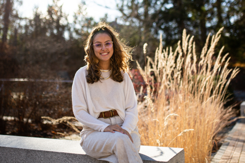A female student smiles, seated on a granite bench. Golden light halos her hair, & shines on a wheat plant to her right. Pine trees blurred in the background.