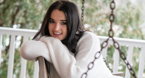 MJ Condon, seated on a porch swing, smiles shyly at the camera. A white fence sits in front of trees in the background, and is out of focus.