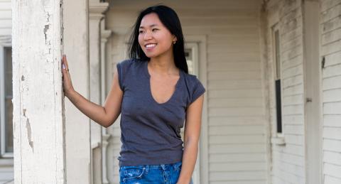 Student smiles, with her head turned to the left, as she places a hand on a column on a front porch. She has long, dark hair, and is dressed casually.