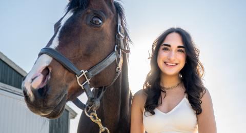 UNH Paul College student Sam Bhacka stands alongside her UNH horse and smiles. The space in between her and her horse is haloed by bright sunlight.