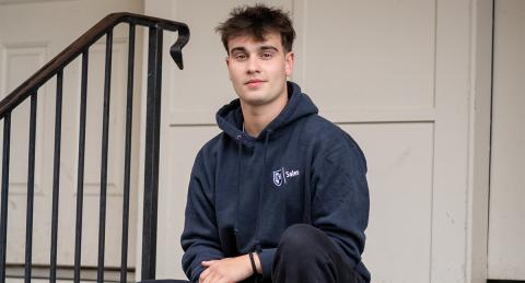 Paul College student sits on a front stoop and gives the camera a small smile. He wears a UNH Sales hoodie. There is a railing to his left, and a door behind.