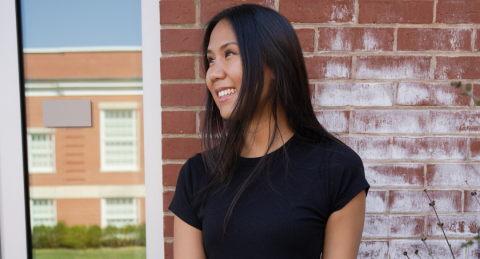 A student stands in front of a brick wall with a window to her left reflecting a  building. She smiles to the the left, and has a black shirt and dark hair.