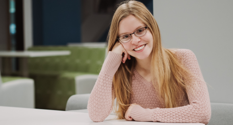 Paul College student smiles, leaning on a table. Booth seating is blurred in the background. She is wearing glasses, a pink sweater, and has strawberry blonde hair.