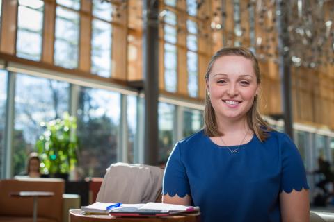 Smiling student Devin McMahon sits with a small desk with a notebook and pen in the foreground. Stippling light streams through windows in the building's atrium.