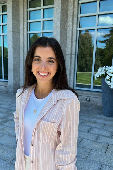 Academic Advisor Julia Payne poses outside with a smile, behind her are three tall windows. She wear a pink linen top and has dark hair.