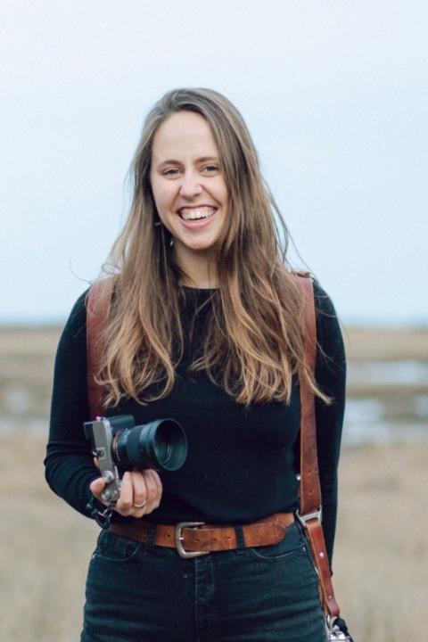 Katherine Keenan headshot. A woman with long brown hair smiling while holding a camera, wearing a black top and jeans with a leather camera strap.