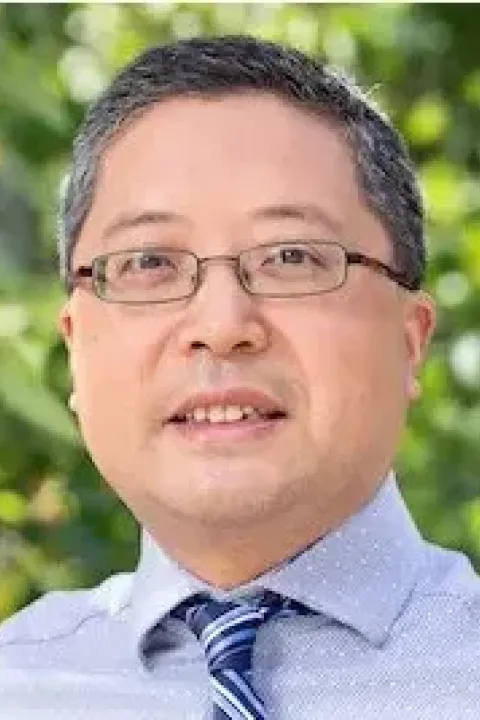 Professional headshot of Jun Li wearing glasses, light blue shirt, and striped tie, smiling against green foliage background.