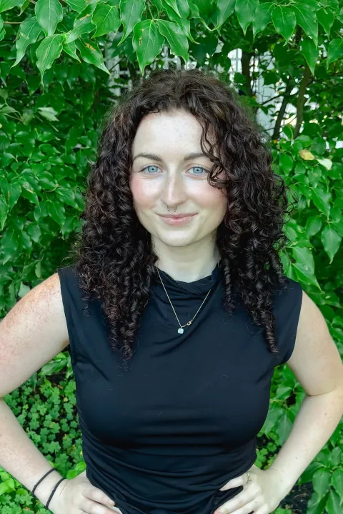 Headshot of Sarah DeSimone; a woman with curly dark hair in black sleeveless top standing against backdrop of green foliage