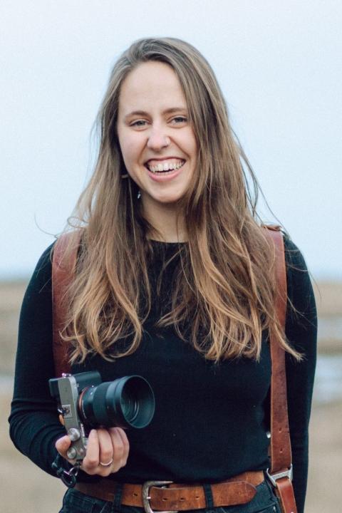Photo of Katherine Keenan standing and holding a camera in a coastal marsh. She has light brown hair and is wearing a brown leather camera harness and a black long sleeve shirt.