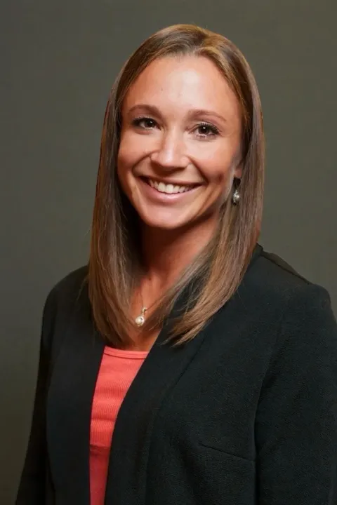 Professional headshot of a Lauren Haley wearing a black blazer over a coral top, with straight shoulder-length hair, against a plain dark background