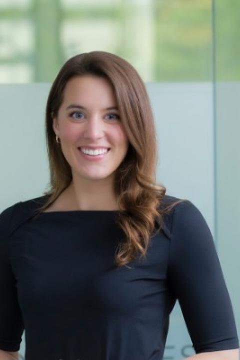 Professional headshot of Alexys Febonio with shoulder-length hair, wearing a black top, smiling, with a softly lit office background.