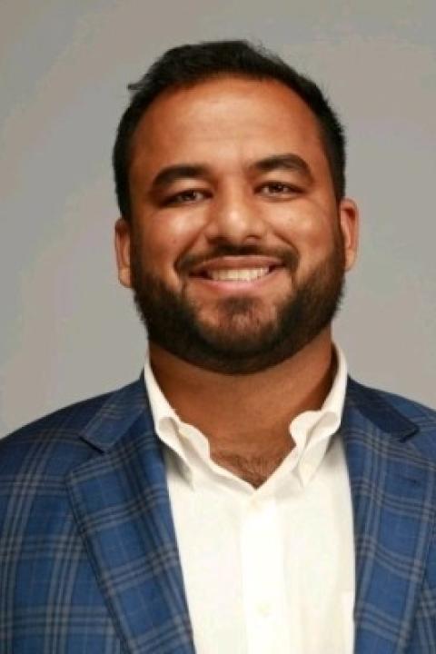 Professional headshot of Hanzla Sheikh with short hair and a beard, wearing a blue plaid blazer and white shirt, against a gray background.