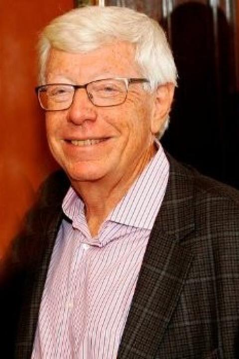 Professional headshot of an Peter Paul with glasses, wearing a patterned blazer and striped shirt, against a warm indoor background.