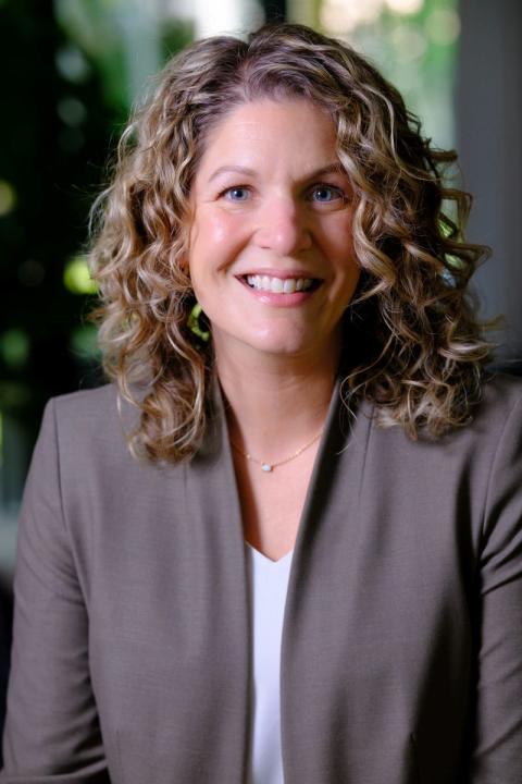 Professional headshot of Suzanne Roberge with curly hair wearing a blazer, seated indoors with a softly blurred background.