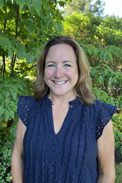 Headshot of a Angela Devoid posing outdoors wearing a navy blouse with green foliage in the background
