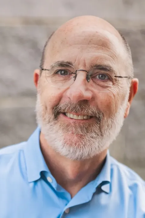 Professional headshot of Jim Gasperoni with glasses and a gray beard wearing a light blue shirt against a blurred stone background.